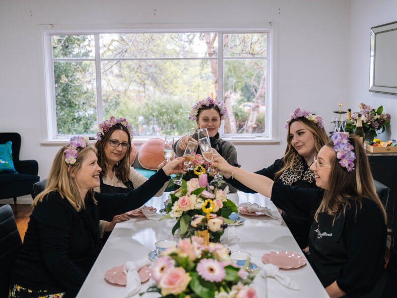A group of women in flower crowns sit around a table, clinking champagne glasses